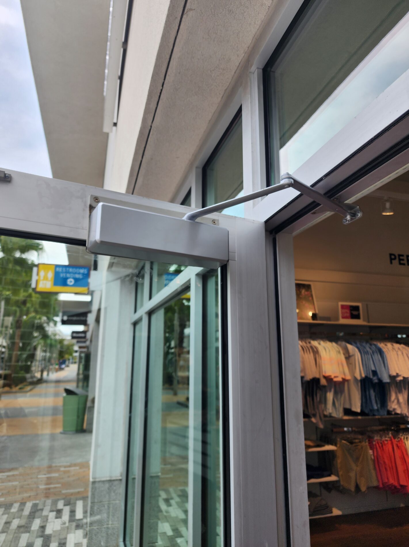 A glass door with a metal arm closer is open. The doorway leads to a clothing store. A city street with a building and a parking sign is visible in the background.