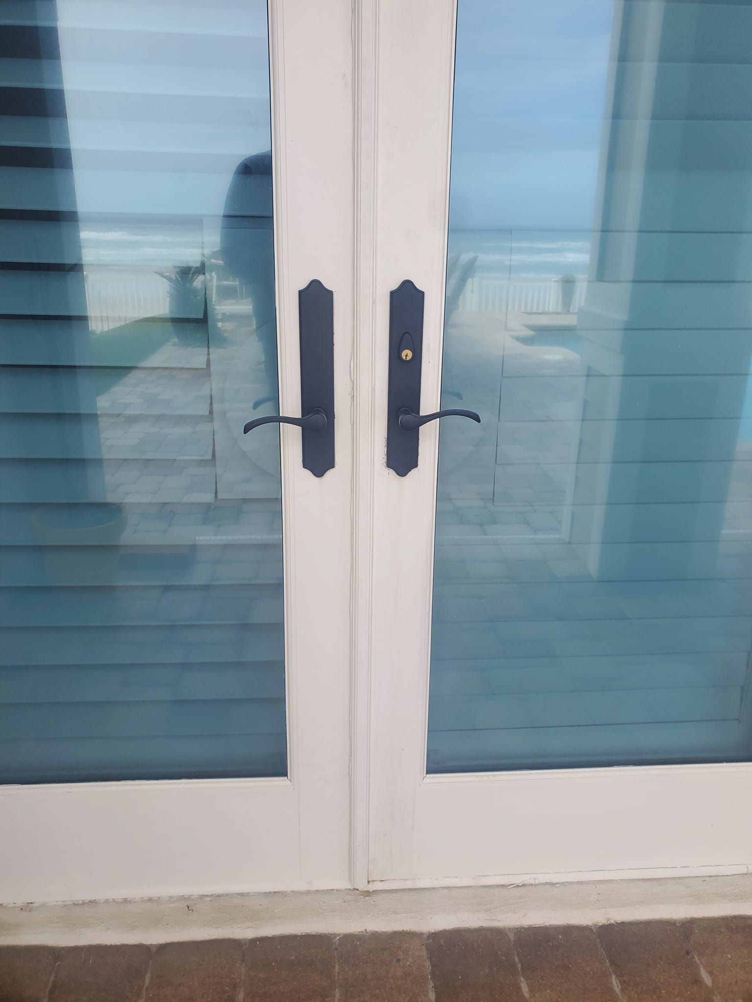 Closed double glass doors with black handles, reflecting an outdoor patio area and ocean view in the background.